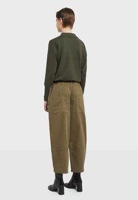 Olivovo zelený pletený polokošeľ s golierom, doplnený o voľné khaki nohavice s dvomi veľkými zadnými vreckami, nosený s čiernymi ihlovými čižmami.