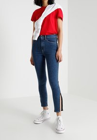 Červené tričko s krátkým rukávem ve spojení s modrými džíny s vysokým pasem a bočními rozparky, nošeno s bílými teniskami. Jednoduchý a neformální design.