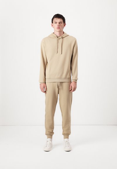Un sweat à capuche beige et un jogging assorti en tissu doux, dotés d'une capuche à cordon, de poignets côtelés et de poches latérales, associés à des baskets blanches.