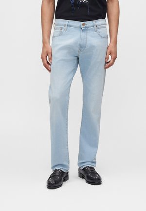 Person trägt hellblaue Jeans mit geradem Schnitt, schwarze Loafers mit Nieten und ein schwarzes Hemd mit blau-weißem grafischem Design.