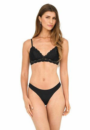 Frau trägt schwarzen Spitzen-Bralette und passende schwarze Tanga-Unterwäsche, steht vor weißem Hintergrund.