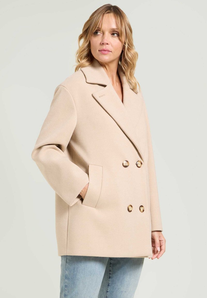 Mujer con cabello rubio ondulado, vistiendo un abrigo beige de doble botonadura con grandes solapas y jeans de color azul claro, de pie frente a un fondo liso.