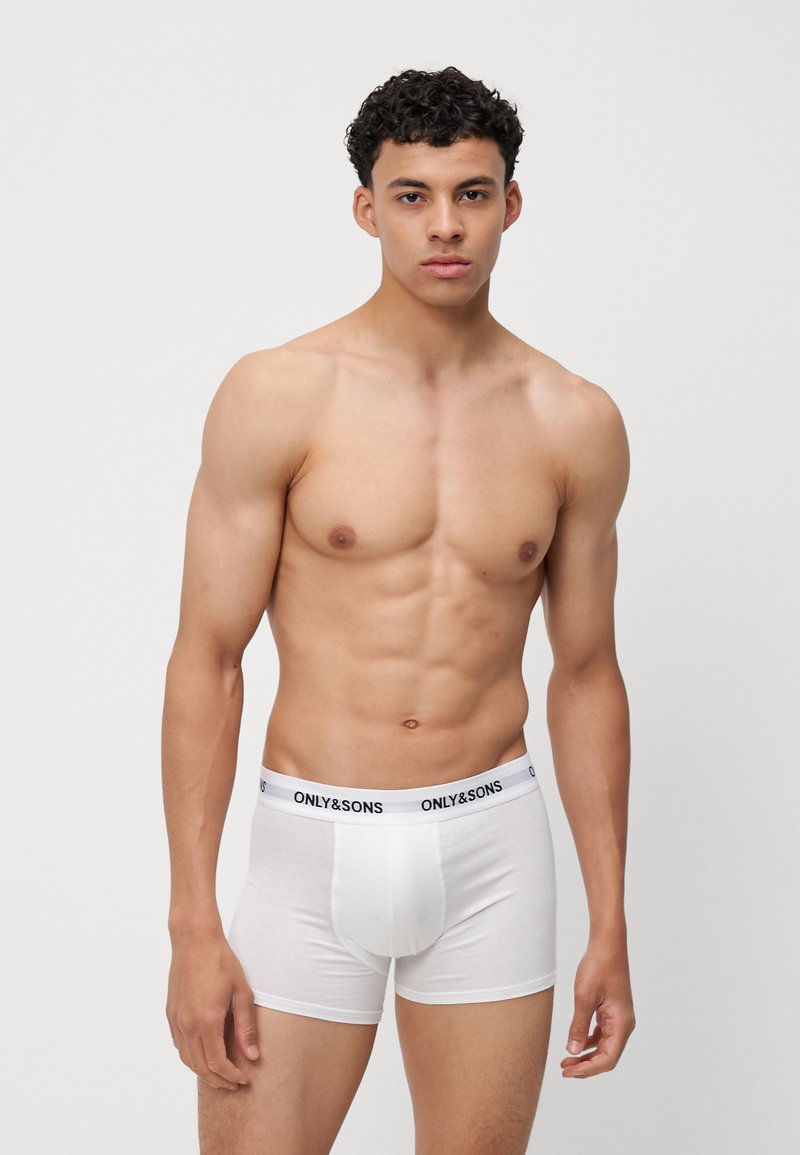 Giovane uomo con capelli ricci che indossa boxer bianchi a marchio Only & Sons, in piedi su uno sfondo grigio chiaro uniforme.