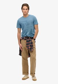 Camiseta de algodón azul con texto en relieve, pantalones de lienzo beige remangados en los tobillos y una camisa de cuadros atada a la cintura. Zapatillas ligeras completan el look.