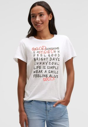 Young woman wearing a white T-shirt with black and red positive text and light blue jeans, smiling with one hand in pocket.