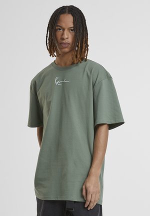 Mlad moški s pletenicami do ramen, ki nosi ohlapno zeleno majico z majhnim belim logotipom na prsih na enobarvnem ozadju.