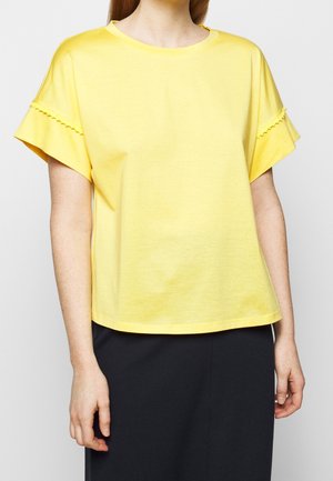 Person iført en løs gul kortærmet top med bølgekant ved ærmerne og et sort skørt mod en ensfarvet baggrund.