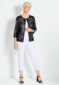 Zwarte neplederen jas met bloemenperforaties, witte top, witte broek met patroon aan de boorden en zwarte hoge hakken.