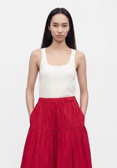 Femme aux longs cheveux foncés portant un haut blanc sans manches et une jupe rouge taille haute froncée avec des poches, debout devant un fond uni.