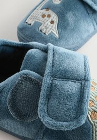 Chaussons en peluche bleus avec une sangle en velcro, ornés de motifs animaliers brodés et d'appliqués dans des teintes bleu clair et marron. Fini en texture douce.