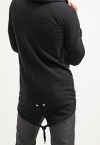 Černá kapucí bunda s prošívanými ramenními akcenty, dlouhým zadním dílem, stahovací pasem a patentními knoflíky na lemu. Hladký povrch látky.