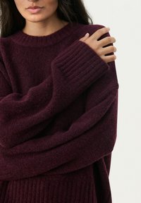 Pullover oversize bordeaux con polsini a costine e una finitura morbida e testurizzata. Presenta un ampio scollo e maniche dalla vestibilità ampia.