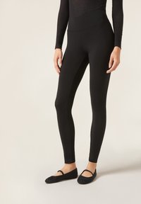 Leggings neri realizzati in un tessuto elasticizzato con una vita alta, caratterizzati da una superficie liscia e un design aderente, abbinati a ballerine nere.