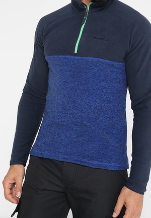 Man wearing a long-sleeve, blue and navy half-zip pullover with black pants, standing against a plain background.