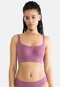 Žena s vrkôčkami má na sebe zladenú mauve bezšvovú textúrovanú podprsenku a nohavičky na jednofarebnom svetlom pozadí.