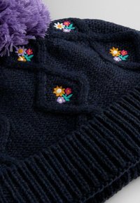 Pletená námořnická čepice s chlupatým fialovým bambulí a květinovou výšivkou v žluté, růžové a bílé barvě. Má vroubkovanou strukturu po obvodu.