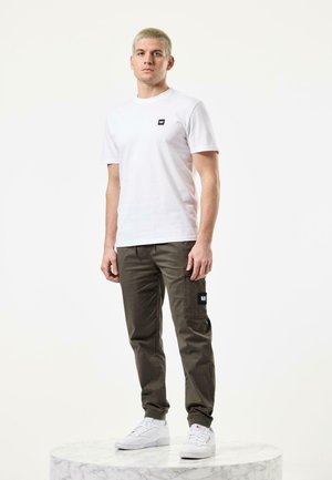 Hvid bomulds-t-shirt med et sort logopatch, parret med olivenfarvede cargo-bukser med en sidelomme og et sort patch. Hvide sneakers fuldender looket.