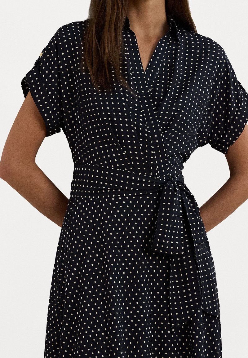 Robe marine à pois blancs, manches courtes retroussées, design enveloppant, taille nouée et silhouette évasée, fabriquée dans un tissu léger.