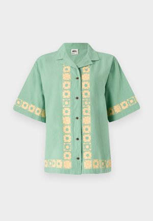 Chemise verte à manches courtes avec col, ornée d'un motif floral jaune sur le devant et les manches. Fermeture à boutons uniforme. Texture douce.