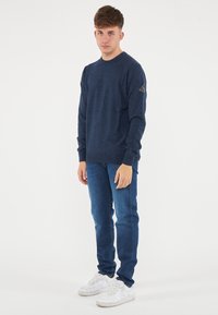 Maglione a maglia blu navy con maniche lunghe e scollo rotondo, abbinato a jeans blu aderenti e sneakers bianche. Presenta un logo sulla manica.