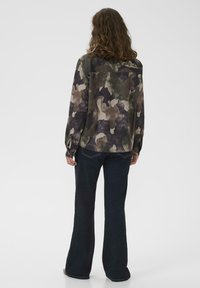 Persona con capelli ricci che indossa una camicia a maniche lunghe in stile camouflage e jeans scuri a zampa d'elefante, in piedi contro uno sfondo chiaro e uniforme.