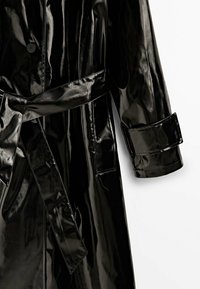 Trench-coat en vinyle noir brillant avec ceinture nouée à la taille et bride au poignet sur une manche longue, sur fond blanc.