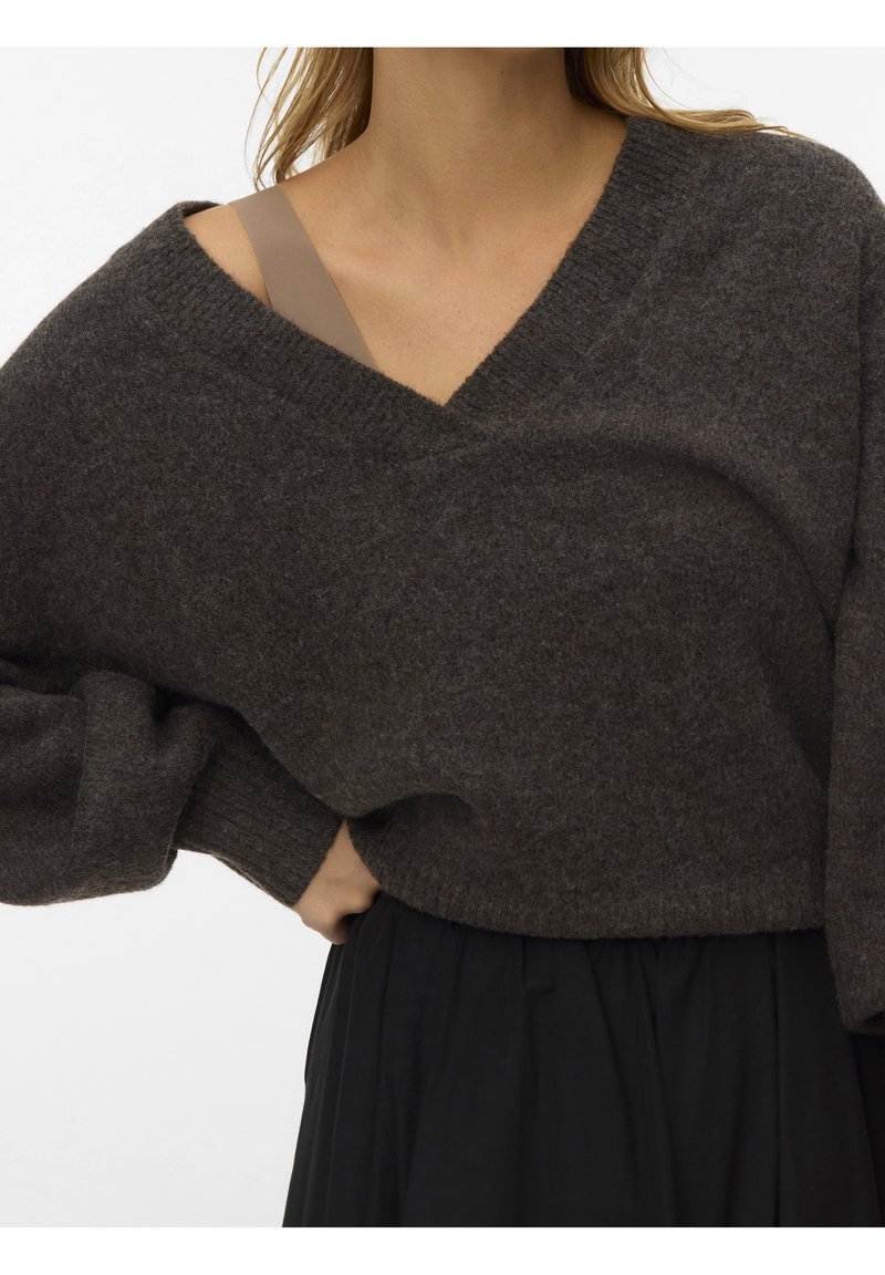Femme portant un pull oversize gris foncé à col en V avec une bretelle de soutien-gorge beige visible et un pantalon noir plissé, main reposant sur sa taille.
