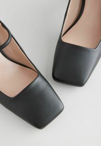 Scarpe in pelle nera con design a punta quadrata, caratterizzate da un'apertura tra la punta e la caviglia e una superficie liscia.
