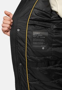 Zwarte pufferjack met gele accentpiping. Beschikt over een ritssluiting zak en een stoffen label met productinformatie. Gelaagd, gewatteerd ontwerp.
