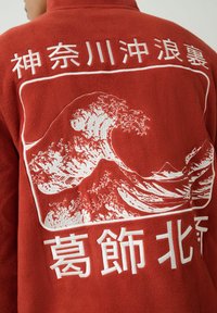 Piros polárdzseki, fehér hímzéssel, amely egy nagy óceáni hullámot, hegyet és japán karaktereket ábrázol a hátoldalon.