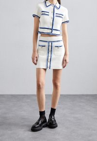 Bílý texturovaný dvoudílný set obsahující zkrácenou bundu s krátkým rukávem a mini sukni, zdobený modrým lemován�ím a knoflíky.