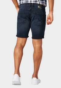 Hombre con pantalones cortos de mezclilla oscuros remangados, zapatillas blancas y una camisa de cuadros metida por dentro, de pie de espaldas sobre un fondo liso.