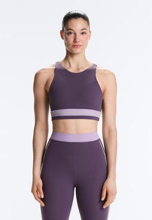 Žena stojaca čelom k prednej časti, oblečená v bezrukávovom fialovom krátkom tričku a fialových legínach s vysokým pásom na jednoduchom svetlom pozadí.