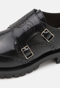 Chaussure en cuir noir à finition brillante, équipée d'une large lanière, de deux boucles métalliques et de détails décoratifs perforés sur le côté.