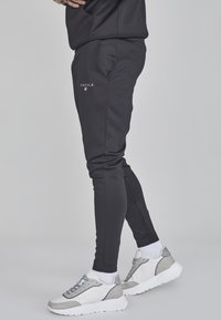 Pantaloni jogger neri aderenti con una superficie liscia, caratterizzati da un logo ricamato sulla coscia sinistra e abbinati a sneakers voluminose bianche e grigie.