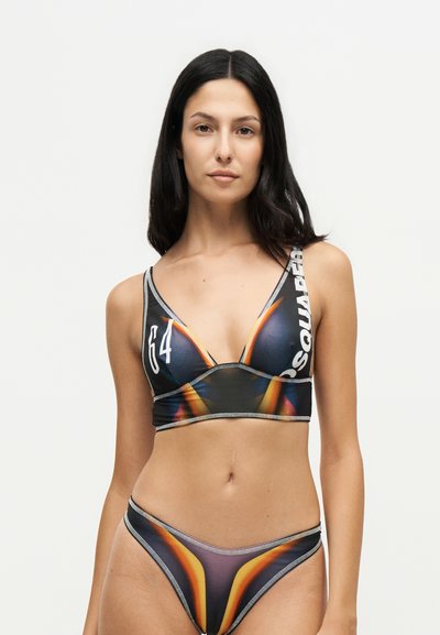 Femme aux longs cheveux foncés portant un bikini noir, orange et jaune à motifs avec un texte blanc sur le haut, sur un fond blanc uni.