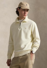 Jeune homme portant une casquette beige, un sweat-shirt crème à fermeture éclair partielle, un pantalon kaki et une montre-bracelet, debout avec une main dans la poche, regardant sur le côté.