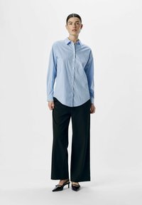 Blauw-wit gestreept overhemd met lange mouwen, gecombineerd met wijde zwarte broek en zwarte puntige schoenen. Model staat in een neutrale omgeving.