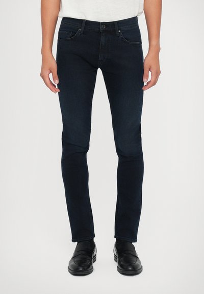 Sötétkék farmernadrág, slim fit szabással és enyhe kopással, öt zsebes dizájnnal és gombos záródással, fekete mokaszinokkal viselve.