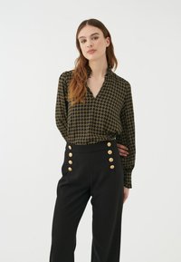 Blusa nera con motivo geometrico dorato, maniche lunghe e scollo a V. Abbinata a pantaloni neri a vita alta con dettagli in oro sui bottoni.