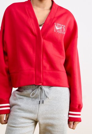Osoba nosząca czerwoną kurtkę Nike na zamek z białymi paskami na mankietach oraz szare spodnie dresowe Nike ze sznurkiem w pasie.