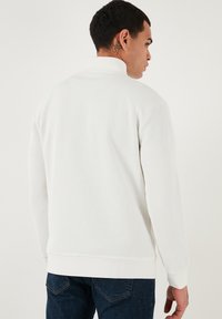 Biely rebrovaný sveter s vysokým golierom a dlhými rukávmi, ktorý má textúrovaný povrch. Skombinovaný s tmavomodrými džínsami.