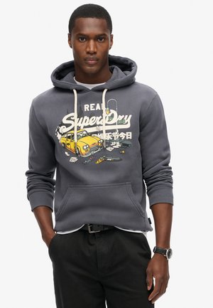 Sudadera de algodón gris con un bolsillo frontal, cordones blancos y un gráfico colorido que presenta un taxi amarillo y elementos de diseño. Pantalones negros y un reloj.