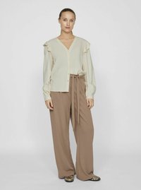 Lys beige skjorte med knapper og flæseærmer, parret med højtaljede brune brede bukser og mønstrede sko. Glat stoftekstur.