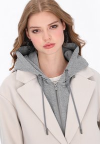 Felpa grigia con zip indossata sotto un cappotto crema, caratterizzata da una texture morbida e coulisse. Il modello indossa grandi orecchini a cerchio neri.