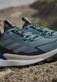 Trail running shoes with a textured teal upper, black accents, and three gray stripes. Features GORE-TEX material and turquoise laces.