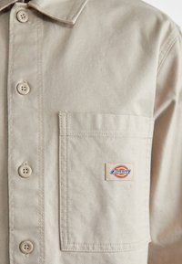 Beżowa koszula zapinana na guziki z kieszonką z przodu, z małą wielokolorową naszywką marki Dickies.