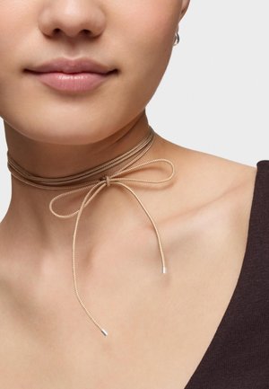 Primo piano del collo di una persona che indossa un sottile choker beige annodato a fiocco, con una maglietta scura e un orecchino visibile.