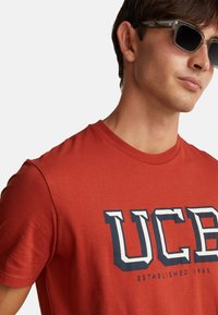 Červené bavlněné tričko s krátkými rukávy, které má velké logo "UCB" v tmavě modré a bílé barvě; hladký materiál a design s kulatým výstřihem.