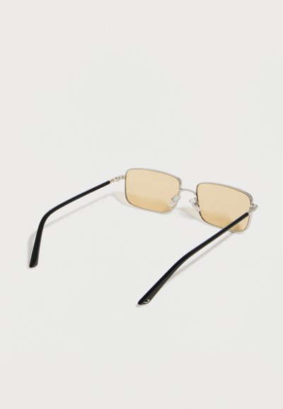 Ορθογώνια γυαλιά ηλίου με ασημένιους μεταλλικούς σκελετούς, φακούς ανοιχτού κεχριμπαριού και μαύρα βραχίωνα ναών, πάνω σε λευκό φόντο.